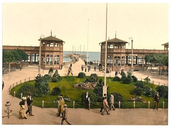 The image depicts a historical scene from the Kurgarten and Landungsbrücke in Zoppot, East Prussia (now part of Sopot, Poland), dating between approximately 1890 to around 1900. The composition shows two wooden pavilions with ornate architectural features such as curved roofs and decorative railings. These structures are situated near a waterfront where people can be seen walking along the pier extending into the sea.

A well-manicured garden area surrounds one of the pavilion, featuring neatly trimmed hedges, flower beds, and trees, indicating an urban park setting. People in early 20th-century attire stroll through the garden or walk along the promenade near the water's edge. Some individuals wear hats indicative of fashion from that era.

In the background, a few sailboats are visible on the horizon, adding to the scenic view of this seaside locale during what appears to be late afternoon based on the lighting and shadows cast by the figures in the scene. The overall atmosphere suggests leisurely activity among residents or visitors enjoying an outing at this picturesque location during that period in German history.