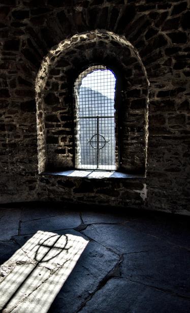 Das Bild zeigt ein vergittertes Fenster in einer Steinmauer. Das Fenster ist bogenförmig und lässt helles Licht herein, das einen Schatten des Gitters auf den Steinboden wirft.