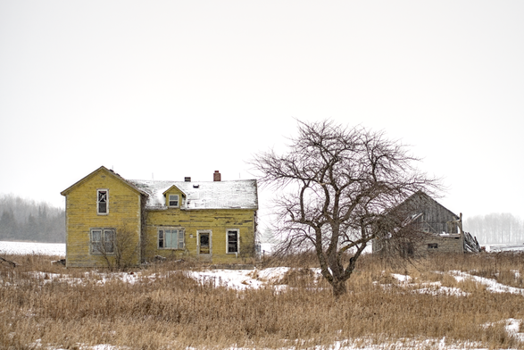 An abandoned yellow wooden farmhouse with chipped paint, broken windows, and a snow-covered roof stands in a snowy rural field. A leafless tree is to the right, with a deteriorating barn partially visible behind it. The background features a hazy treeline and a pale, overcast sky.