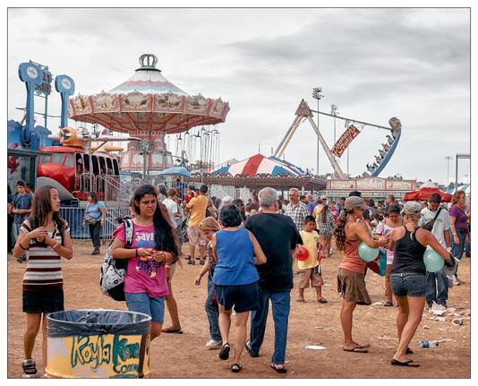 This image depicts a lively scene at a local fair or carnival. There are a variety of amusement rides visible, including a traditional carousel and a large pendulum ride. The area is bustling with people of different ages and backgrounds walking around, enjoying snacks, and interacting with one another. The ground appears to be a dirt surface, indicating the event might be taking place on a temporary setup or in an open field. The sky is slightly overcast, giving the scene a soft, diffuse lighting. Several booths and attractions with colorful decorations and signs such as "Popcorn", "Cotton Candy", and "Mini Donuts" suggest the availability of typical fair concessions. This image captures the casual, festive atmosphere commonly found at such events.