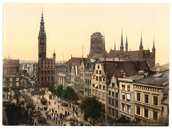 This historical image depicts a bustling urban scene in Danzig, also known as Gdańsk. The central focus is the Langen Market and Court House with its notable architecture featuring spires and large clock towers. Surrounding buildings show various architectural styles from the period, including brick facades, dormer windows, and ornate detailing around doors and balconies.

In the foreground, a crowd of people can be seen gathered in what appears to be an organized procession or event on Market Square, with individuals dressed in military-style uniforms marching. The atmosphere is lively yet orderly, indicative of community gatherings typical during that era's social events.

The overall color palette consists of muted sepia tones, giving the photograph an aged and historical feel.
