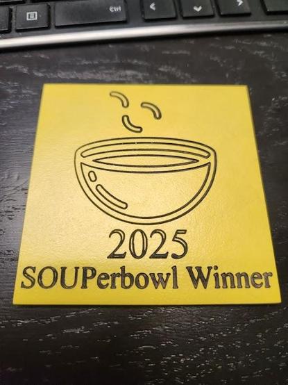 A yellow square with a soup bowl engraved on it. Below the bowl is text reading "2025 SOUPerbowl Winner". Superbowl is spelled s o u p.