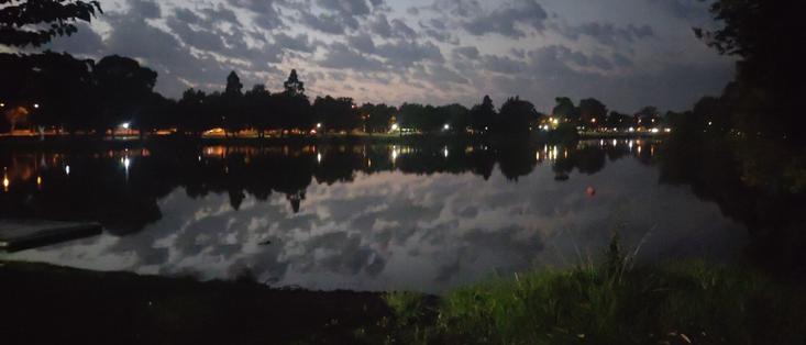 A predawn photo at lake weeroona. Scattered clouds in the dark orange to light blue sky are reflected in the still lake. Dark trees surround, with car and town lights on the distance