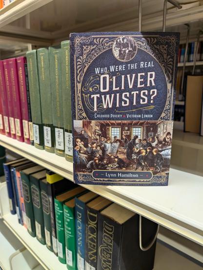 The book "Who Were the Real Oliver Twists?" by Lynn Hamilton, positioned on the Dickens bookshelf of our library. Behind and below the book are many more books on and by Dickens visible