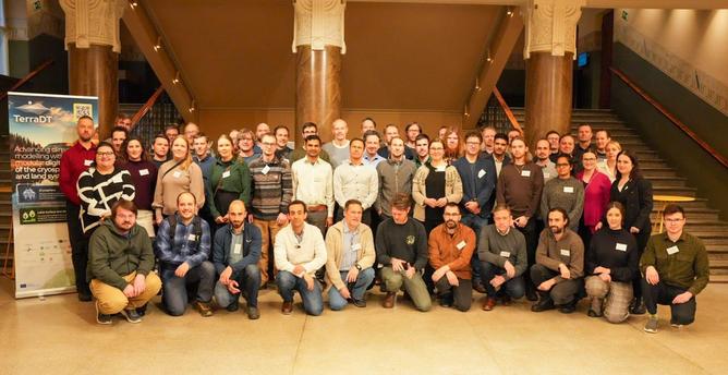 The TerraDT project team in a foyer in front of three massive pillars, a roll-up on the left side says "TerraDT". On both sides, an upward-leading stairwell is visible.