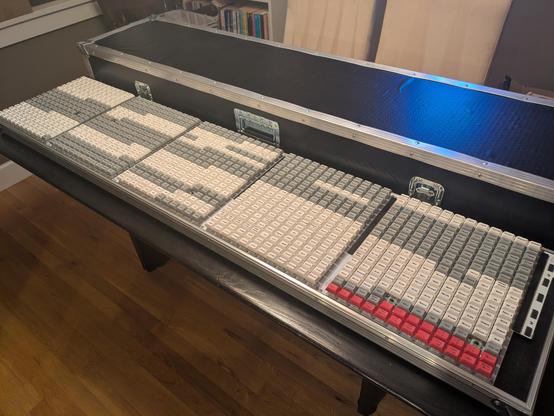 A massive, ridiculous, utterly impractical keyboard that is 60 columns wide and 17 deep, split into 5 sub-panels. Each key is one of the 1000 most common English words, with the keycaps alternating between white and grey by letter. At the bottom right there is also a section of red keys. The keyboard is built into the bottom of a road case, with the lid in classic black laminated plywood and extruded aluminum edges sitting behind it.