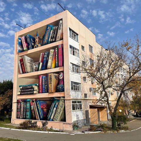 A stunning 3D mural by JanIsDeMan in Solnechnodolsk, Russia, transforms the side of a residential building into an enormous bookshelf. The hyper-realistic trompe-l'œil artwork features several shelves filled with detailed books, each displaying vibrant spines with Cyrillic titles, alongside a small Russian nesting doll and a miniature architectural model. The mural seamlessly integrates into the building's facade, creating an illusion that the bookshelves are built into the structure itself. A breathtaking celebration of literature and street art.