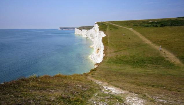 A scenic view of a white chalk cliff coastline bordering a calm blue sea, with a grassy hill and path in the foreground.