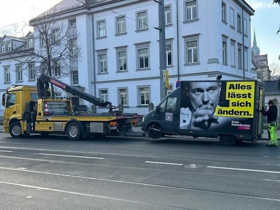 Ein gelbes Abschleppfahrzeug hebt ein schwarzes Lieferfahrzeug an, das mit einem Wahlplakat der FDP versehen ist. Auf dem Plakat steht „Alles lässt sich ändern“ neben einem Porträt von Christian Lindner. Im Hintergrund sieht man ein weißes Gebäude mit mehreren Fenstern und eine ruhige Straßenumgebung. Ein Arbeiter in Warnkleidung steht neben dem Lieferfahrzeug.
