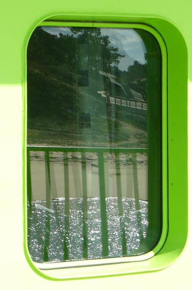 A imagem mostra uma vigia do ferry que faz a travessia do rio Sado, entre Setúbal e Tróia. A moldura da vigia é verde brilhante e a forma retangular com cantos arredondados. A vigia reflete uma área de praia fluvial com uma onda cinza escura e inúmeros reflexos de luz a desvanecer-se na areia molhada e uma encosta coberta de vegetação e arvoredo verde-escuro. Em primeiro plano está o gradeamento escuro da amurada e interior da embarcação. No topo vislumbra-se um céu azul-pálido nublado.