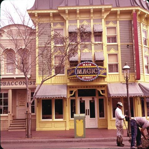 A colorful two-story building features a yellow façade with a sign that reads "Main Magic Street, Jokes & Tricks." Two individuals are seen working in front near a trash can, and a tree is partially visible.