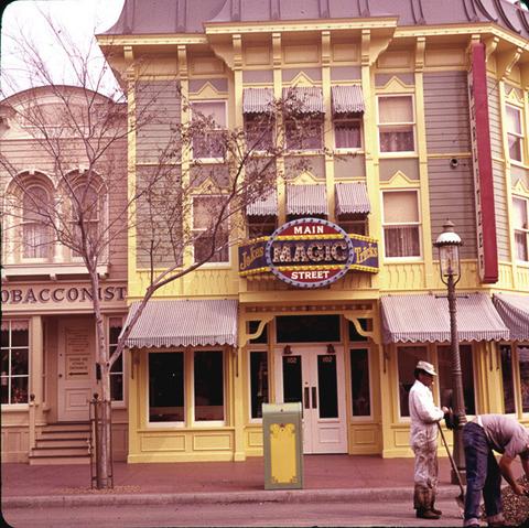 A colorful two-story building features a yellow façade with a sign that reads "Main Magic Street, Jokes & Tricks." Two individuals are seen working in front near a trash can, and a tree is partially visible.