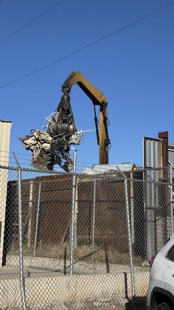 Heavy crane with claw lifting scrap metal, behind a metal fence, then a wooden fence, framed against a bright blue sky.
