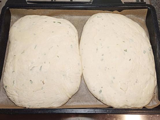 Zwei große, ungebackene Fladenbrote liegen auf einem mit Backpapier ausgelegten Backblech. Der helle, aufgegangene Teig enthält kleine grüne Kräuterstückchen, die gleichmäßig verteilt sind. Die Oberfläche ist leicht uneben, mit einigen sichtbaren Lufteinschlüssen. Das Blech steht auf einem Herd oder einer Arbeitsfläche in einer Küche.