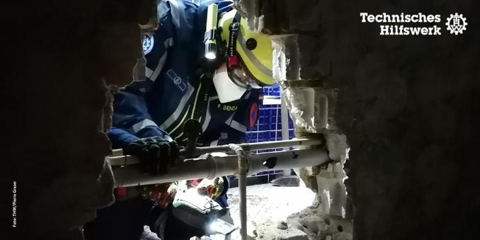 Durch eine Stück herausgebrochener Mauer hindurch ist eine Person in Einsatzkleidung des Technischen Hilfswerks gezeigt, die mit einer Säge die Mauersteine bearbeitet. Sie trägt einen gelben Helm und eine Atemschutzmaske. Im Hintergrund sind blaue Fließen.