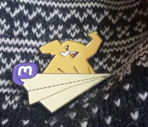 Pin of a happy yellow Mastodon who sits on a paper plane. A white M on a purple square with rounded corners forms the tail of the plane. The merch pin is stuck on a Norwegian sweater.