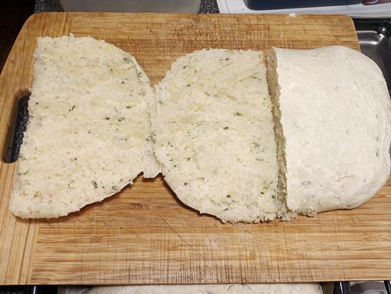Ein frisch gebackenes Fladenbrot liegt auf einem Holzschneidebrett. Es wurde aufgeschnitten, sodass die weiche, luftige Krume mit kleinen Kräuterstückchen sichtbar ist. Die Kruste ist hell und eher weich, nicht stark gebräunt. Das Brot hat eine lockere, leicht feuchte Textur. Ein weiteres Stück liegt ungeschnitten daneben. Das Schneidebrett zeigt leichte Gebrauchsspuren und liegt auf einer Küchenarbeitsfläche.