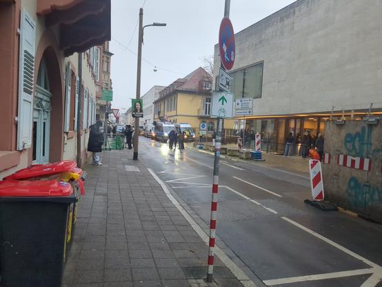 Strasse vor dem Eingang zum Bundesgerichtshof in Karlsruhe. Rechts ein klotzartiges Gebäude, davor eine nasse Strasse, links zwei Mülltonnen. Ein paar Polizisten