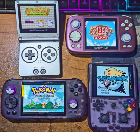 A bunch of retro emulation handheld consoles on a desk. One looks like a silver Gameboy Advance SP. Another is a 1:1 pinkish purple handheld. Another one is a horizontal atomic purple handheld. And the last one looks like an atomic purple Gameboy Color but with extra buttons and joysticks.