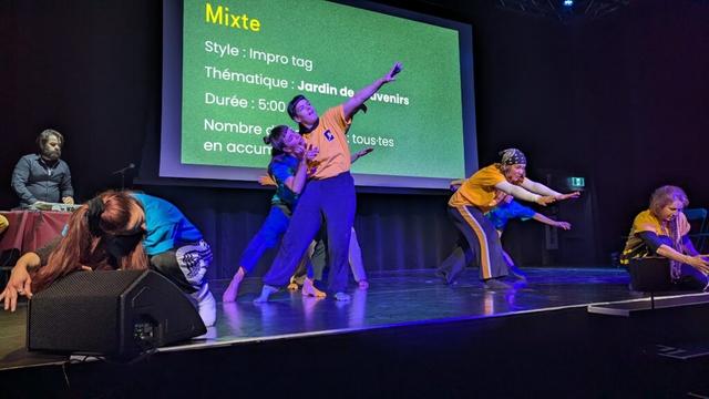 Huit danseuses professionnelles se sont affrontées lors d’un match d’impro-danse fin janvier à la bibliothèque Gabrielle-Roy. Au centre, les artistes Nelly Paquentin (à gauche) et Julia-Maude Cloutier. Une équipe de danse porte des gilets jaunes, l'autre, des gilets verts. La scène et le fond de scène sont noirs. Un écran de projection montre les informations sur l'improvisation en cours, sur fond vert.  (Photo : Alice Zanetta)