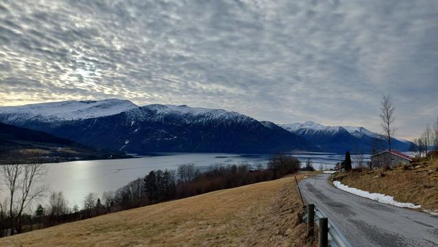 A fjord going from left to right. In the foreground, a yellow-brown field to the left and a narrow asphalt road to the right, both sloping downwards. On the other side of the fjord, tall dark blue mountains with snow on the tops. The sky is tightly dotted with small clouds. The sunlight is coming through the clouds to the left.