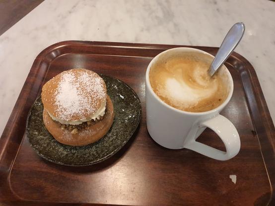 Tummalla tarjottimella laskiaispulla, pistaasitäyte näkyy tummana juovana kermavaahdon alta. Oikealla puolella iso kahvikuppi, jossa cappuccinoa.