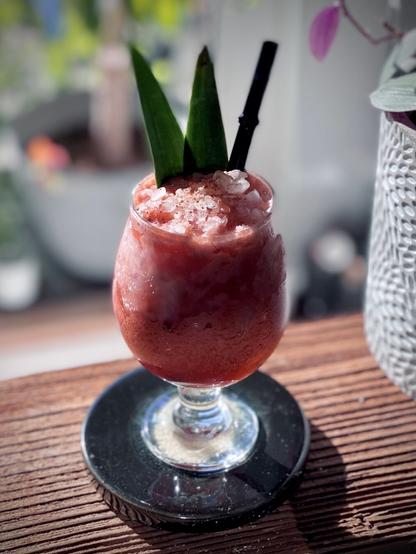 pink blended cocktail with pineapple frond garnish sitting on a wooden shelf with the sun shining