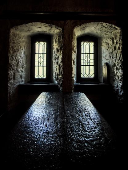 Das Bild zeigt einen dunklen Raum mit zwei Fenstern, durch die Licht eindringt. Das Licht beleuchtet einen dunklen, möglicherweise hölzernen Tisch oder eine Oberfläche. Die Wände um die Fenster herum scheinen aus Stein zu sein und haben eine raue Textur. Der Raum ist insgesamt düster und geheimnisvoll.