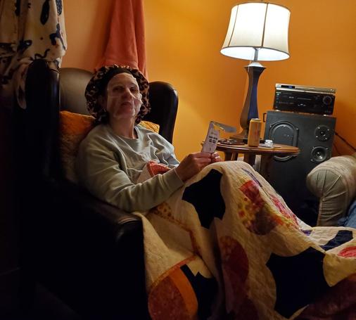 Sarah, relaxes in a comfortable armchair, wearing a facial mask and a leopard-print shower cap. She is draped in a colorful quilt and holds a product packaging in her hand. The background features an orange wall, a lamp turned on, and a small side table with a stereo system and various items.