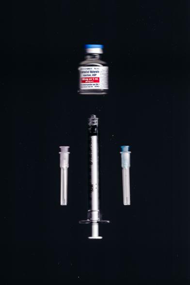 A composition featuring a vial of Estradiol Valerate, a syringe, and two needle caps arranged symmetrically against a black background. The objects appear slightly blurred, creating a sense of movement or distortion.