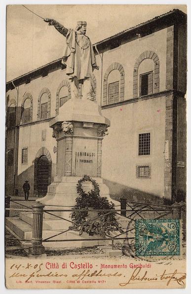 The image displays a vintage postcard featuring an aged photograph of the Garibaldi Monument in Città di Castello, Italy. The monument depicts General Giuseppe Garibaldi standing on a pedestal with his right hand extended upwards holding what appears to be a sword or pole. His posture suggests leadership and determination.
In the background stands a building with several arched windows and classical architectural elements such as pilasters and ornate cornices. There is a person walking in front of the monument, which adds perspective and scale to the scene. The postcard includes handwritten text on its border and corner indicating it was sent from "Città di Castello" (City of Città di Castello) with an address or recipient's name partially legible.
The overall tone of the photograph is sepia-toned, which gives it a historical feel reflective of early photography. The image has been slightly tilted and appears to be scanned at a lower resolution than its original size.