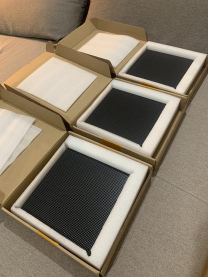 Three black LED Matrix panels in the cardboard boxes I received them in. They are protected by white foam. The boxes are laying on a sofa.