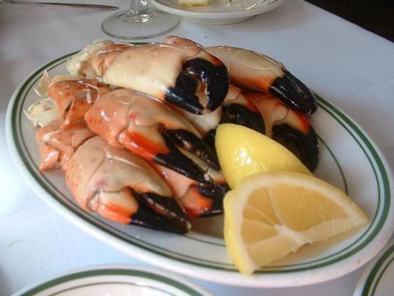 Ein Teller mit mehreren "Florida Stone Crab" Scheren, die neben einer halben Zitrone angerichtet sind. Der Teller hat einen grünen Rand und steht auf einem weißen Tisch. Im Hintergrund sind verschwommen verschiedene Gegenstände zu erkennen.