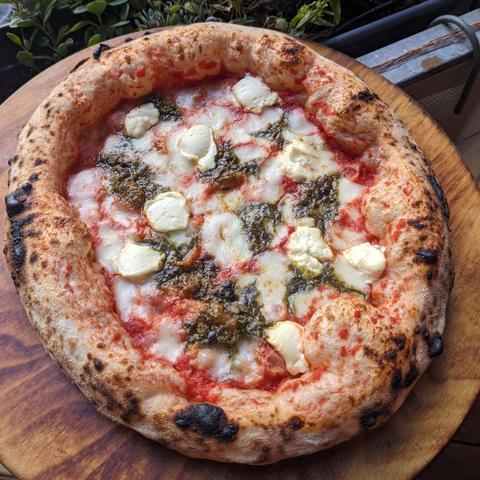 A Pizza Napoletana on a wooden pizza board. It got a big and airy crust and is topped with tomato sauce, basil, Fior di Latte, Mascarpone and Pesto di Pistacchi.