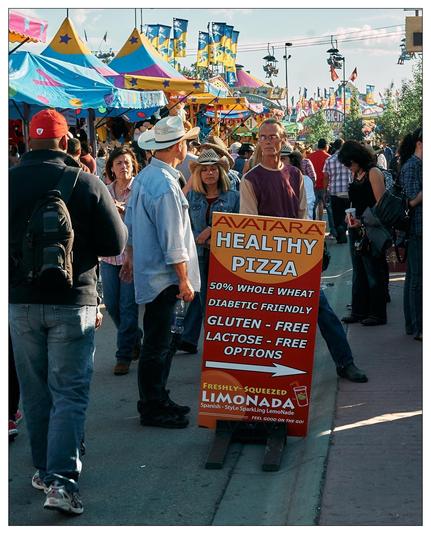 The image captures a lively scene at a fair or festival. In the foreground, you can see a sign advertising "Avatara Healthy Pizza," which mentions several dietary options such as 50% whole wheat, diabetic-friendly, gluten-free, and lactose-free options. Additionally, the sign advertises freshly squeezed, Spanish-style sparkling lemonade referred to as "Limonada."

The background is bustling and colorful with various vendor stalls marked by vibrant flags and decorations. People of different ages are walking around or standing and talking, suggesting a communal and festive atmosphere. Some are dressed casually whereas others, like the two individuals in cowboy hats, have a more distinctive style, which might hint at a local cultural flavor or a themed event at the fair. The setting appears to be outdoors in good weather, contributing to the high turnout and dynamic environment of the event.