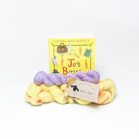 Picture shows a sock set of yarn consisting of a main skein which is yellow with red and purple speckles and a mini skein which is lilac. They are tied together with gold ribbon. Behind the yarn stood on its end is a copy of Jo's Boys by Louisa May Alcott. The book is yellow with purple and red drawn images on it.
