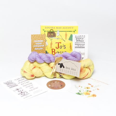 Picture shows a sock set of yarn consisting of a main skein which is yellow with red and purple speckles and a mini skein which is lilac. They are tied together with gold ribbon. Behind the yarn stood on its end is a copy of Jo's Boys by Louisa May Alcott. The book is yellow with purple and red drawn images on it. In front of the book is two bookmarks and in front of the yarn is two postcards and a sticker.
