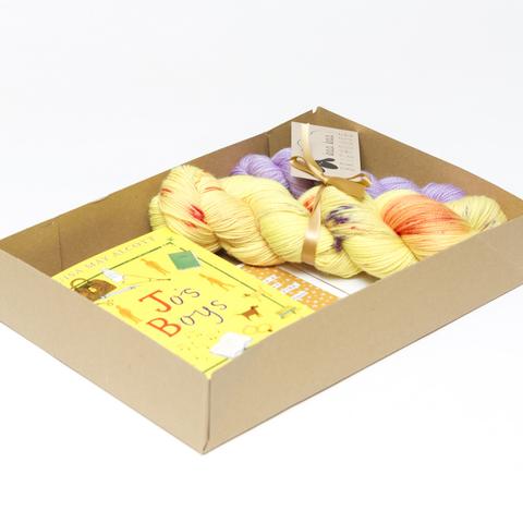 Picture shows an open A4 box containing a sock set of yarn consisting of a main skein which is yellow with red and purple speckles and a mini skein which is lilac. They are tied together with gold ribbon. Beside the yarn is a copy of Jo's Boys by Louisa May Alcott. The book is yellow with purple and red drawn images on it.