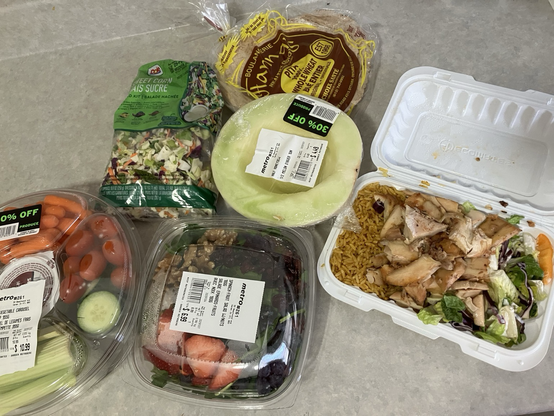 An array of discounted foods including a veggie tray, a fresh spinach and walnut salad, a bagged mixed green salad and a small platter of freshly made shawarma fixings . And half a honeydew melon and pita bread.