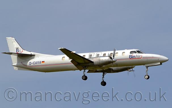Side view of a long, thin, twin propellor-engined airliner flying from left to right at a very low altitude with flaps extending from the rear of the wings and the undercarriage lowered, suggesting it is about to land.
There are thin red and blue stripes running along the body, blue "BinAir" titles on the forward fuselage under the cockpit, and the registration "D-CAVA" on the rear fuselage, also in blue.
Blue-grey sky fills the rest of the frame.