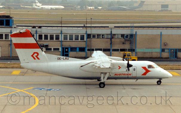 Side view of a white, high-winged, twin propellor-engined airliner taxiing from left to right.
The plane has a stylised red arrowhead on the forward fuselage, just below and aft of the cockpit, and red "Austrian" titles on the lower fuselage.
The same arrowhead is on the white engine pods, in a slightly smaller form. There are 2 red stripes at the top of the tail, and a red stylised upper-case letter "R" at the base, with the registration "OE-LRU"  at the bottom in black.
Grey taxiway fills the foreground, with a 2-storey sandy brown and grey building stretching across the frame in the background.
In the distance, acres of grass lead up to cargo planes in front of hangars  at the top of the frame.