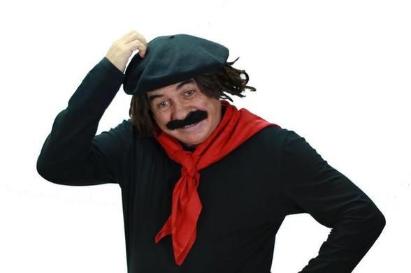 Foto de Guri de Uruguaiana (Jair Kobe) coçando a cabeça. É um personagem de comediante branco fantasiado de gaúcho vestido de preto e lenço vermelho amarrado no pescoço, com bigodão preto postiço e boina preta.