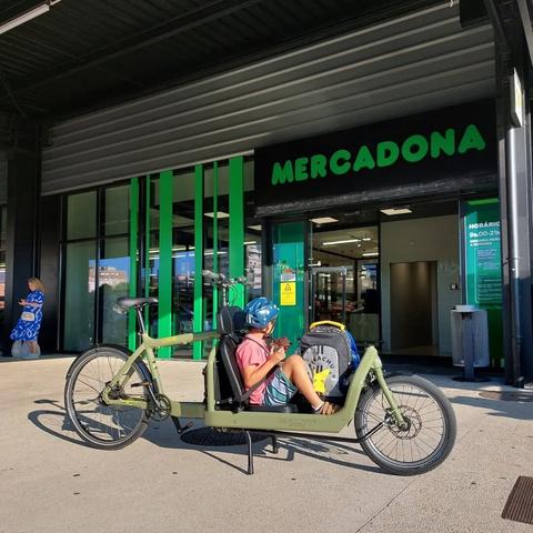Criança dentro de uma bicicleta de carga à frente verde à entrada do supermercado Mercadona
