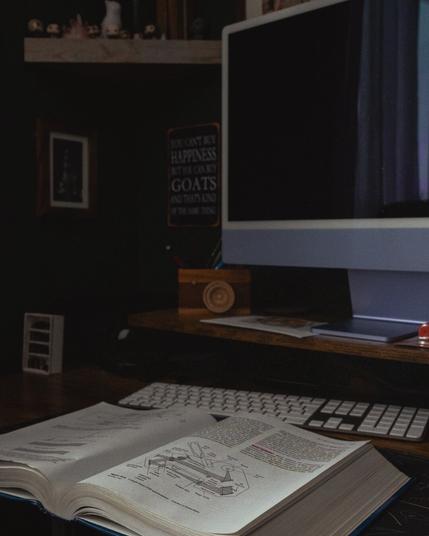 An open textbook on a wooden desk, displaying a detailed technical diagram with labeled components. The desk holds a white keyboard and an iMac with a dark screen. In the background, shelves with small figurines and a framed picture are visible, along with a sign that humorously reads, “You can’t buy happiness, but you can buy goats, and that’s kind of the same thing.” The room has a cozy, dimly lit atmosphere, suggesting a late-night or deep-focus study session.