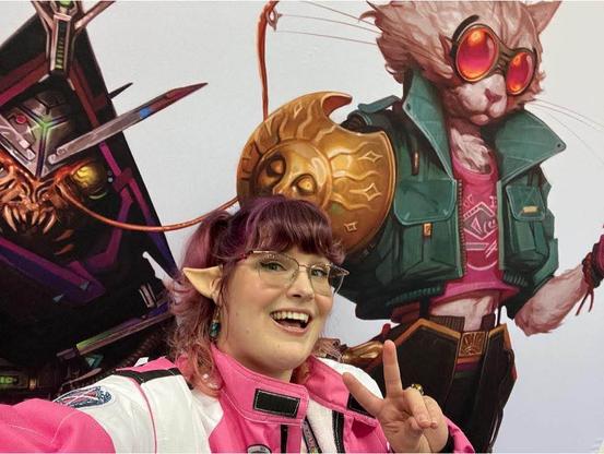 Jenny Jarzabski, a white woman with purple hair, wearing glasses, elf ears, and a pink and white jacket, showing a peace sign in front of artwork of a humanoid cat wearing red goggles and a green jacket over a red shirt and carrying a shield.