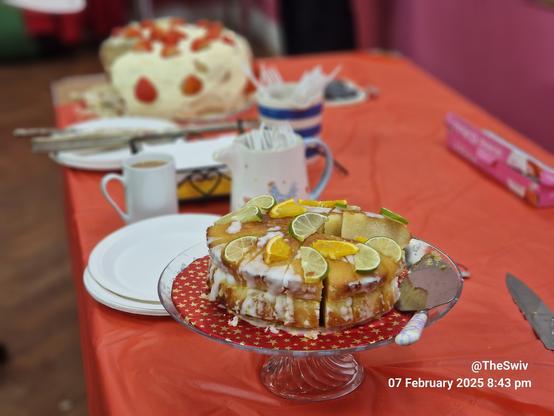 The image shows a red tableclothed table.
In the foreground a lemon and lime drizzle cake. With an out of focus strawberry cream cake at the other end of the table at the top of the image