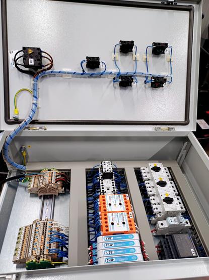 The image shows the inside of a wired control cabinet.

On the inner side of the door are the contacts for push buttons and signal lights, neatly wired and secured.

Inside the cabinet, terminal blocks, motor protection switches, airflow monitors, zener barriers, and a 24V DC power supply are organized and connected on DIN rails.
