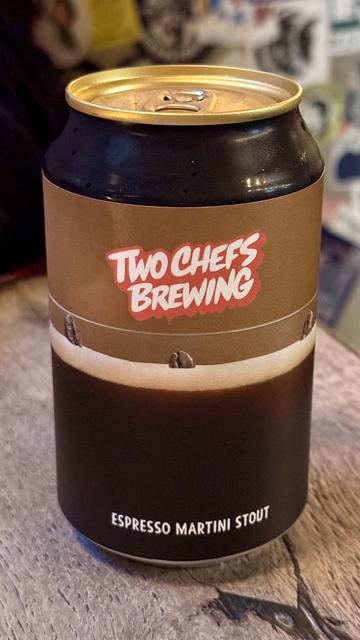 A can of Espresso Martini Stout from Two Chefs Brewing, featuring a dark design with a prominent logo and coffee bean illustrations. The can is placed on a wooden surface.