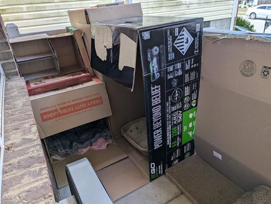 a large electric lawnmower box containing a cardboard litterbox, pizza boxes stacked on top of a Home Depot box, a box fan and a cardboard scratcher