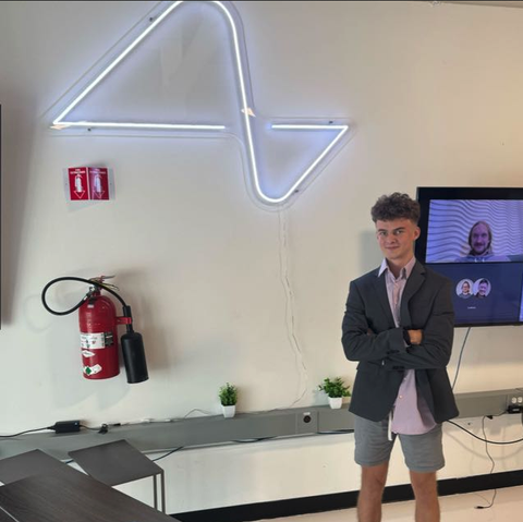 Edward Coristine, seen standing in front of a desk and a TV, wearing grey shorts, a pink shirt and a dark blazer. A neon insignia hangs on the wall. This image was taken from Coristine's WhatsApp profile.